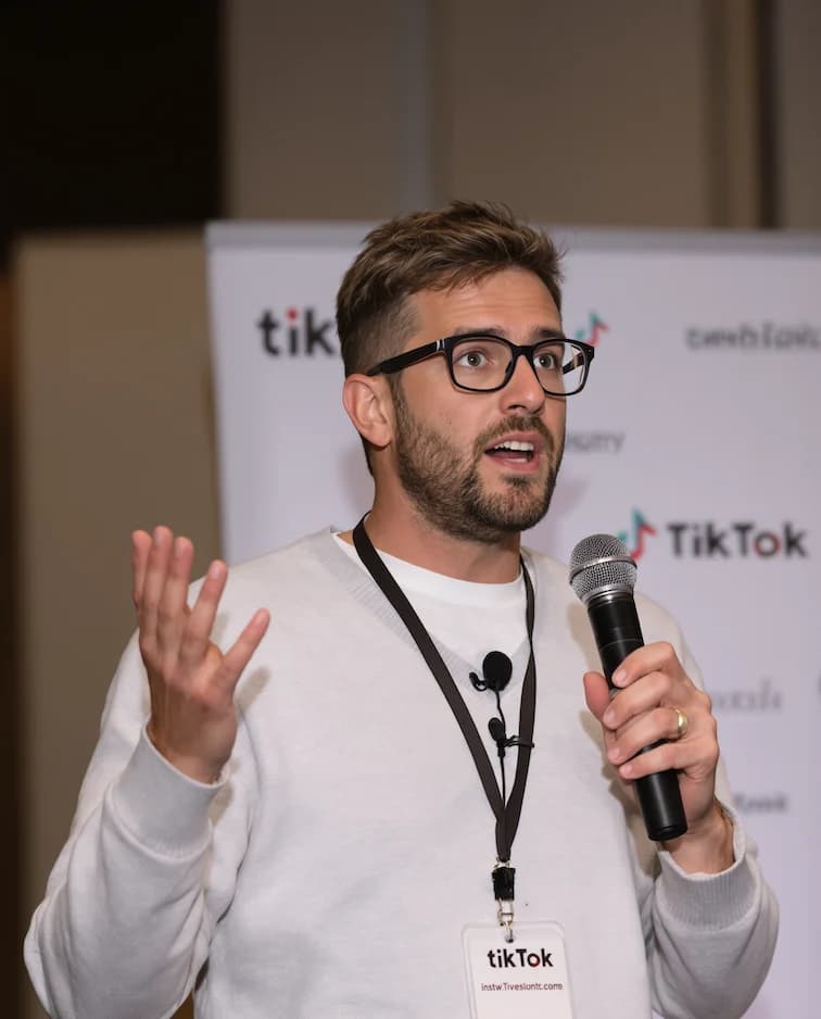 GenImg AI prompt A charismatic speaker is captured mid-speech. He has short, tousled brown hair that's slightly messy on top. He has a round circle face, clean shaven, adorned with rounded rectangular-framed glasses with dark rims, is animated as he gestures with his left hand. He is holding a black microphone in his right hand, speaking passionately.The man is wearing a light grey sweater over a white t-shirt. He's also wearing a simple black lanyard hanging around his neck. The lanyard badge has the text TikTok .Behind him, there is a blurred background with a white banner containing logos and text (including tiktok), a professional conference setting.
